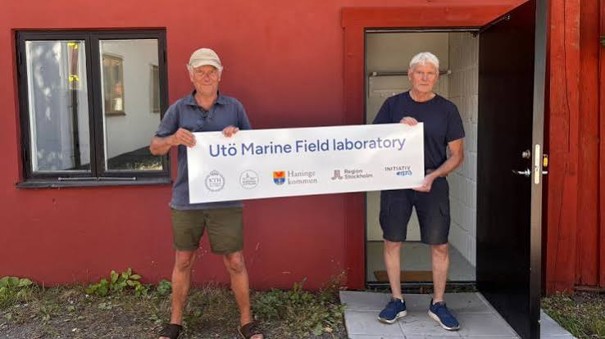 Two men standing outside a red building holding a banner that reads 'Utö Marine Field Laboratory'.